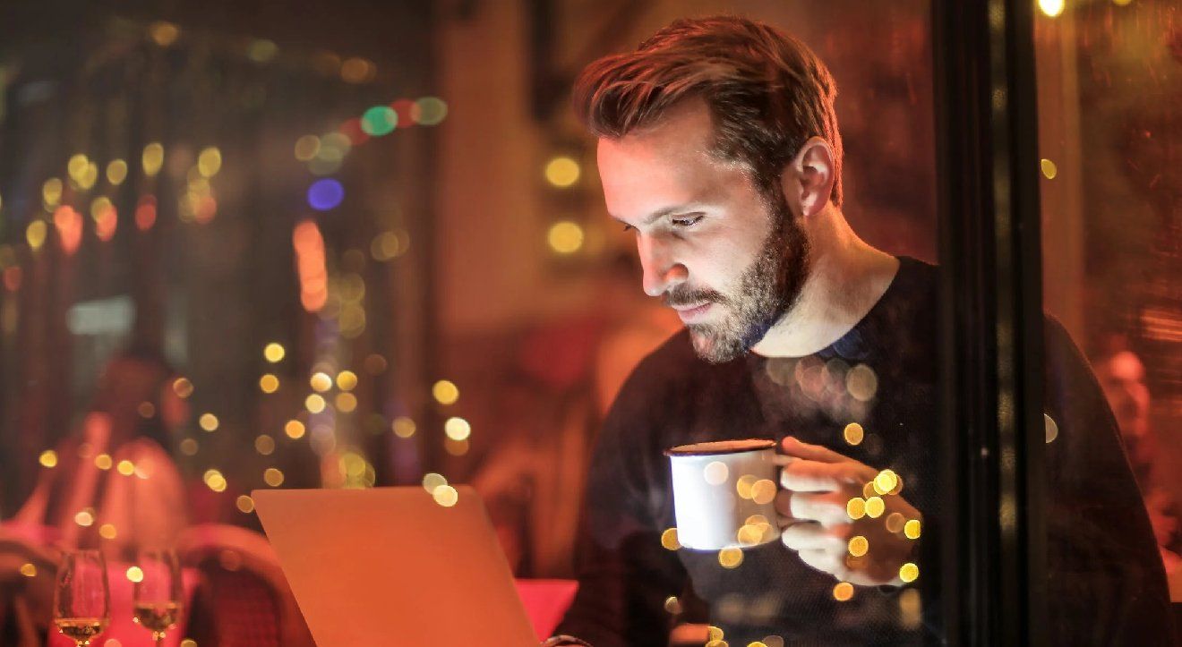 A man drinking a coffee in a dimly lit cafe in front of a laptop.